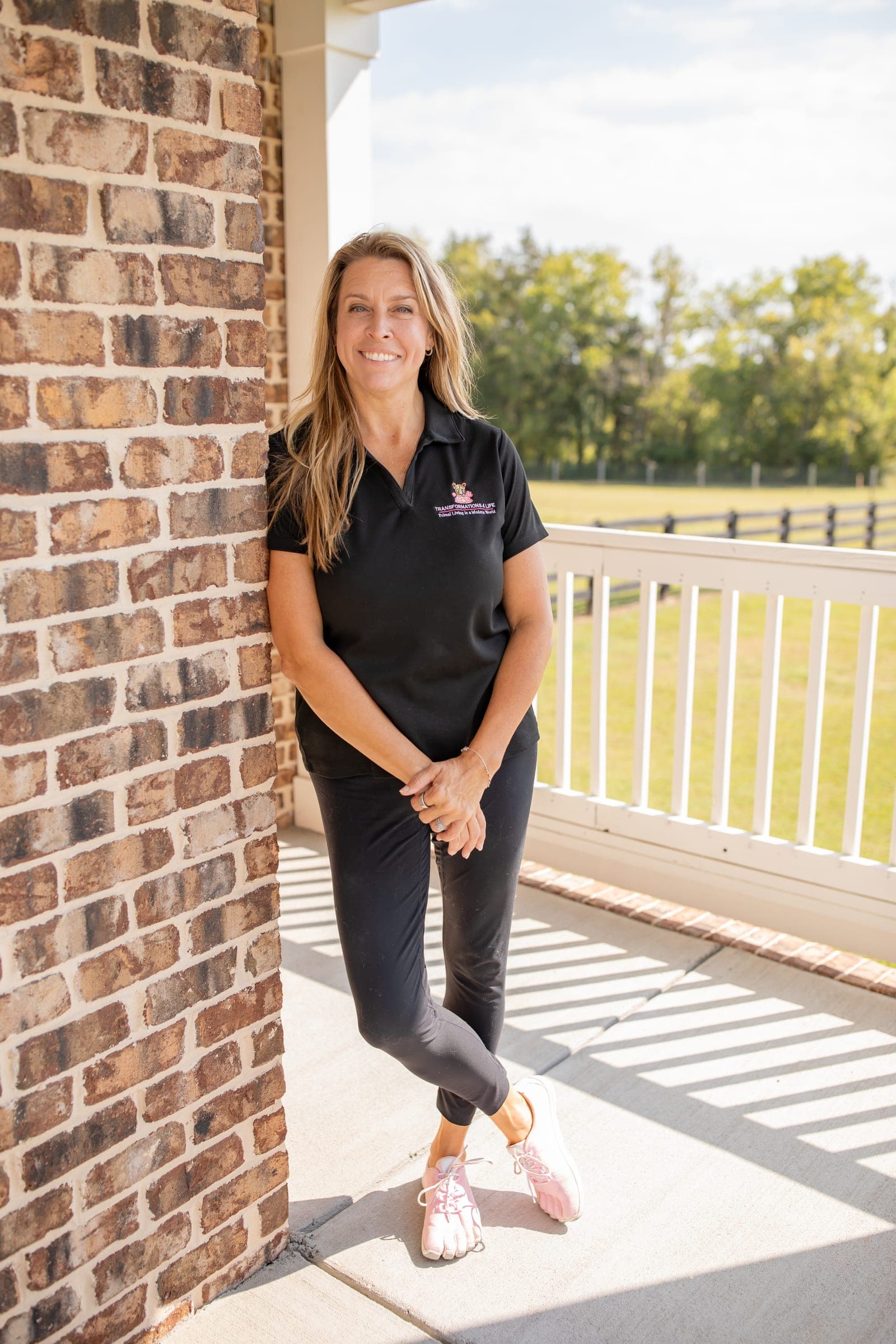 Julie Wilcoxson standing against a brick pillar, ankles crossed and hands clasped, in a black Transformations 4 Life polo and pink barefoot toe shoes.