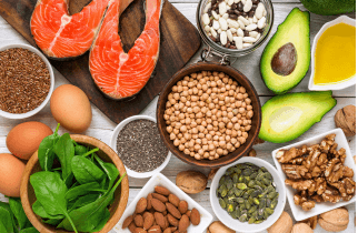 Salmon fillets, eggs, spinach, avocado, walnuts, almonds, pumpkin seeds, chia, flaxseed, chickpeas and olive oil laid out on a pale wooden table.