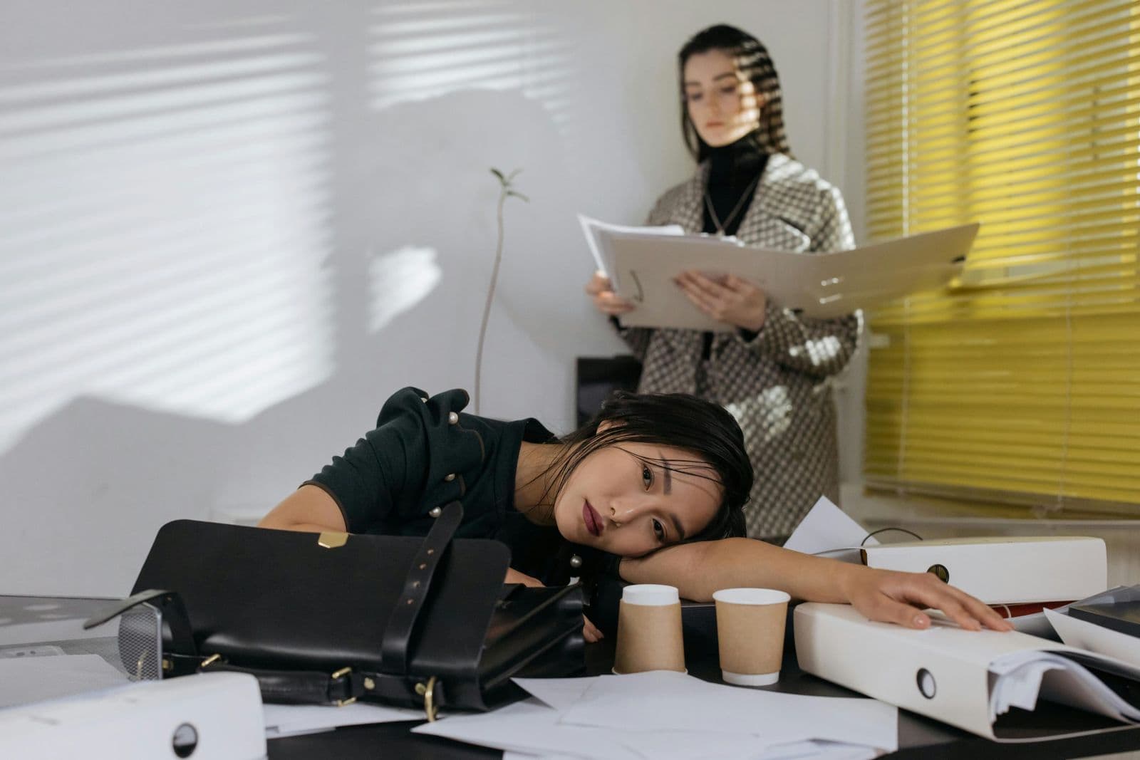 A woman resting her head on a desk buried in paperwork and empty coffee cups, a colleague reading a file behind her.