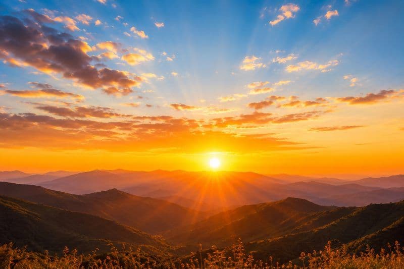 The sun sitting low over layered mountain ridges at sunrise, rays fanning out through orange cloud.